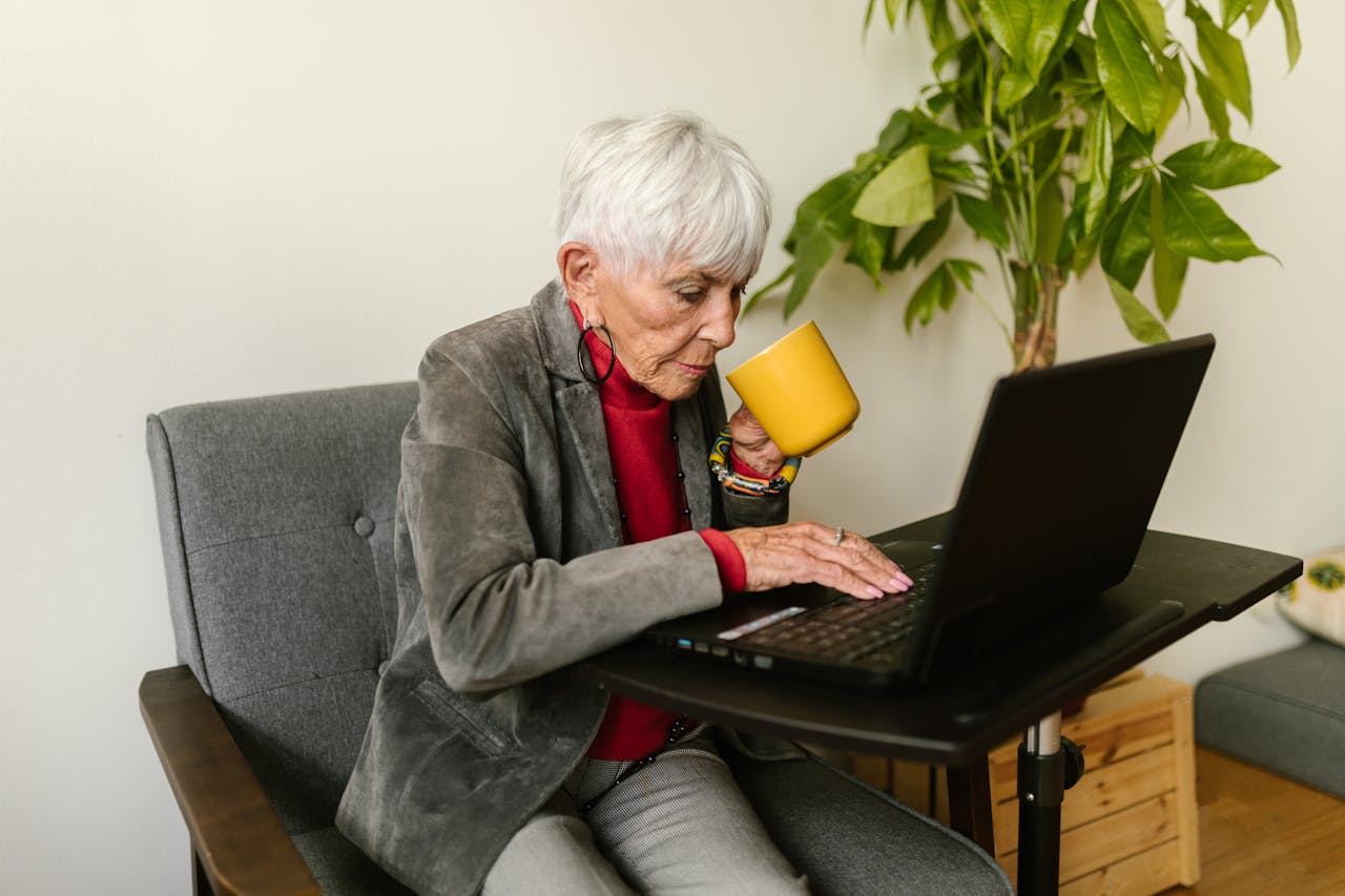 services-04 Elderly woman in gray blazer using laptop while drinking coffee, sitting at a home office desk.
